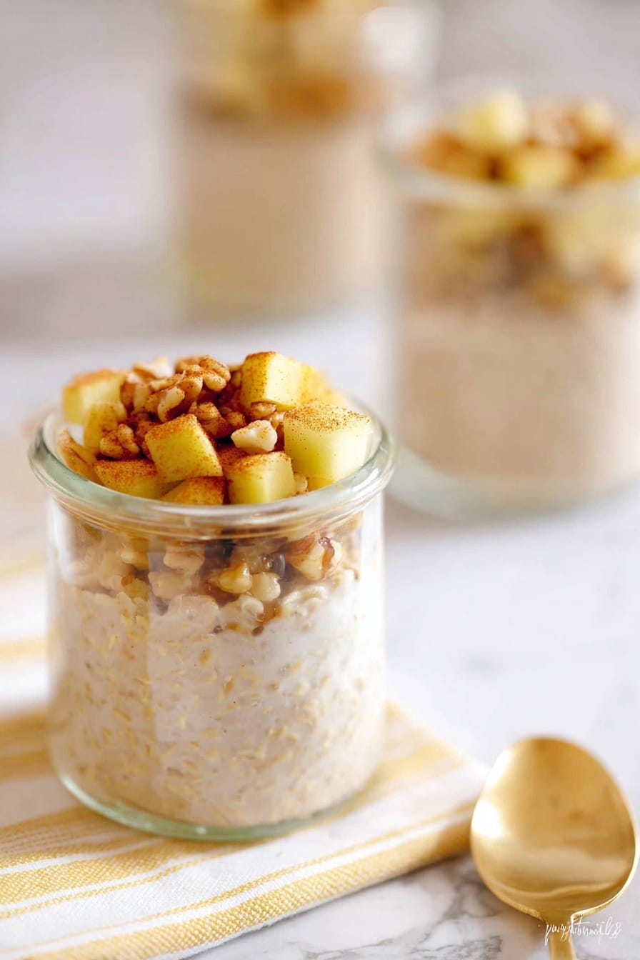A small clear glass jar filled with two layers is shown on a white marbled surface. The bottom layer is creamy oatmeal with a soft texture and light beige color. On top, there is a layer of small, yellowish apple cubes mixed with nuts, all sprinkled with cinnamon, adding a warm brown touch. Another similar jar is blurred in the background. Nearby, a golden spoon rests on a white cloth with thin, yellow stripes. The photo is taken with an iphone --ar 2:3 --v 7 - Apple Cinnamon Overnight Oats, healthy overnight oats recipes, easy breakfast ideas, no-fuss healthy breakfast, cozy oatmeal recipes