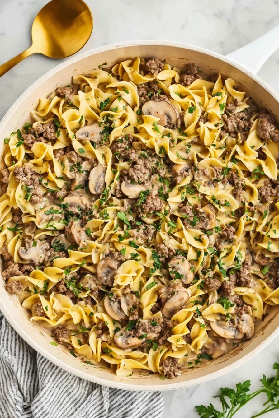 The image shows a wide white pan filled with creamy egg noodles mixed with sliced mushrooms and small pieces of cooked ground beef. The noodles are soft yellow with a smooth texture, spread evenly across the pan. The mushrooms, pale brown and slightly shiny, are mixed throughout the noodles. The beef is browned and scattered on top and inside the noodles in small chunks. Fresh green herbs are sprinkled over the whole dish, adding a pop of color. The pan sits on a white marble surface, next to a gold spoon and white cloth with gray stripes. Photo taken with an iphone --ar 2:3 --v 7 - One-Pot Ground Beef Stroganoff, easy ground beef dinner, creamy beef stroganoff, quick weeknight meals, hearty beef recipes