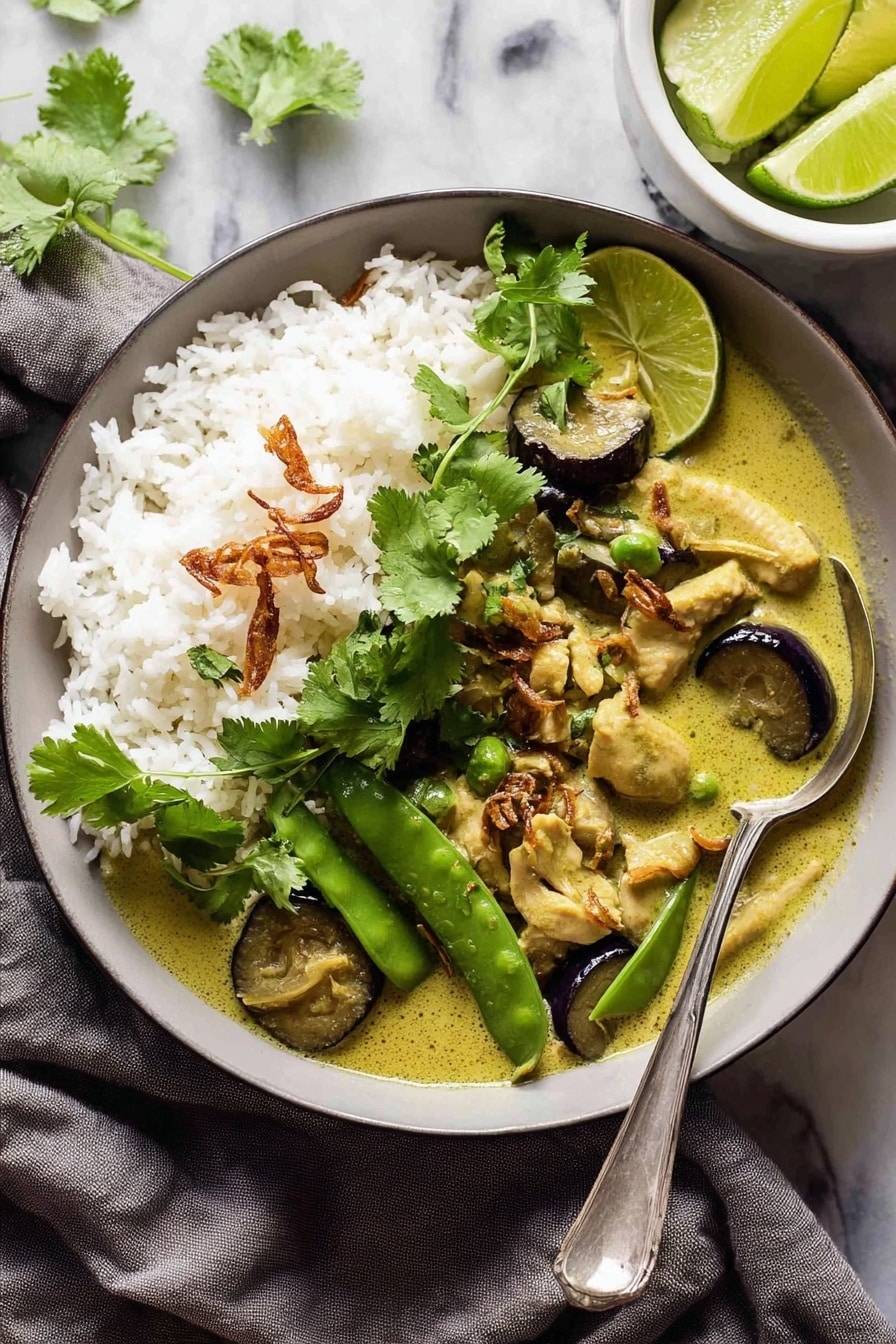 The image shows a black bowl filled with a creamy green curry dish. On the left side, there is a mound of white rice with a few crispy fried shallots sprinkled on top and a small bunch of fresh green cilantro leaves as garnish. The curry has thick slices of dark purple eggplant and fresh green snap peas floating in the light green sauce. A spoon is partially submerged in the curry on the right side of the bowl. The bowl is placed on a soft gray cloth with a small white plate with lime wedges and cilantro at the top right corner, all set on a white marbled surface. Photo taken with an iphone --ar 2:3 --v 7 - Easy Thai Green Curry with Chicken, Thai green curry recipe, quick chicken curry Thai, authentic Thai green curry, spicy green curry with chicken