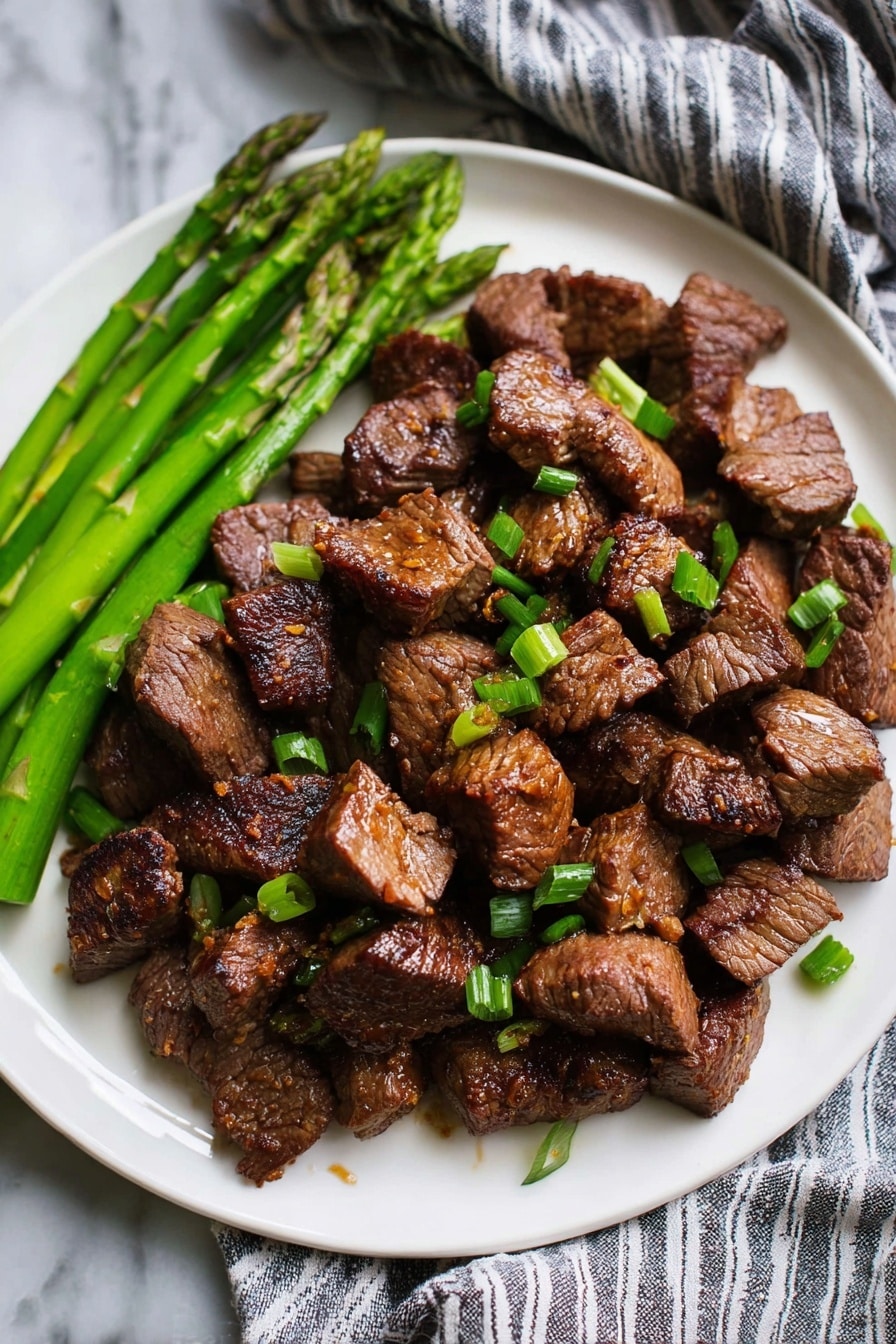 The image shows a white plate filled mostly with many small brown pieces of cooked beef chunks piled together in the center. The beef has a slightly shiny texture with caramelized edges and is garnished with small bright green chopped scallions scattered on top. On the left side of the plate, there are several long, bright green asparagus stalks lying next to the beef. The plate rests on a cloth with gray and white stripes, and the background is a white marbled texture. Photo taken with an iphone --ar 2:3 --v 7 - Garlic Honey Steak Bites, Garlic Honey Steak Bites Recipe, Easy Steak Bites, Quick Steak Dinner Ideas, Flavorful Steak Snacks