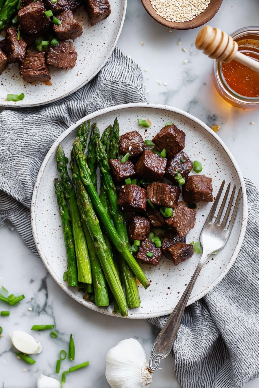 The dish shows a white speckled plate with two layers: on the left side, there are bright green asparagus spears laid out neatly, while on the right side, there are dark brown, seared beef cubes scattered with small pieces of chopped green onions on top and around them. A silver fork rests on the right edge of the plate. The background is a white marbled surface with a gray and white striped cloth partially under the plate, and there is another similar plate with the same food partly visible in the top left corner. A jar of honey with a wooden dipper and a small bowl with white sesame seeds sit on the surface above the plate, and at the bottom left are scattered green onion pieces and a white garlic bulb. Photo taken with an iphone --ar 2:3 --v 7 - Garlic Honey Steak Bites, Garlic Honey Steak Bites Recipe, Easy Steak Bites, Quick Steak Dinner Ideas, Flavorful Steak Snacks