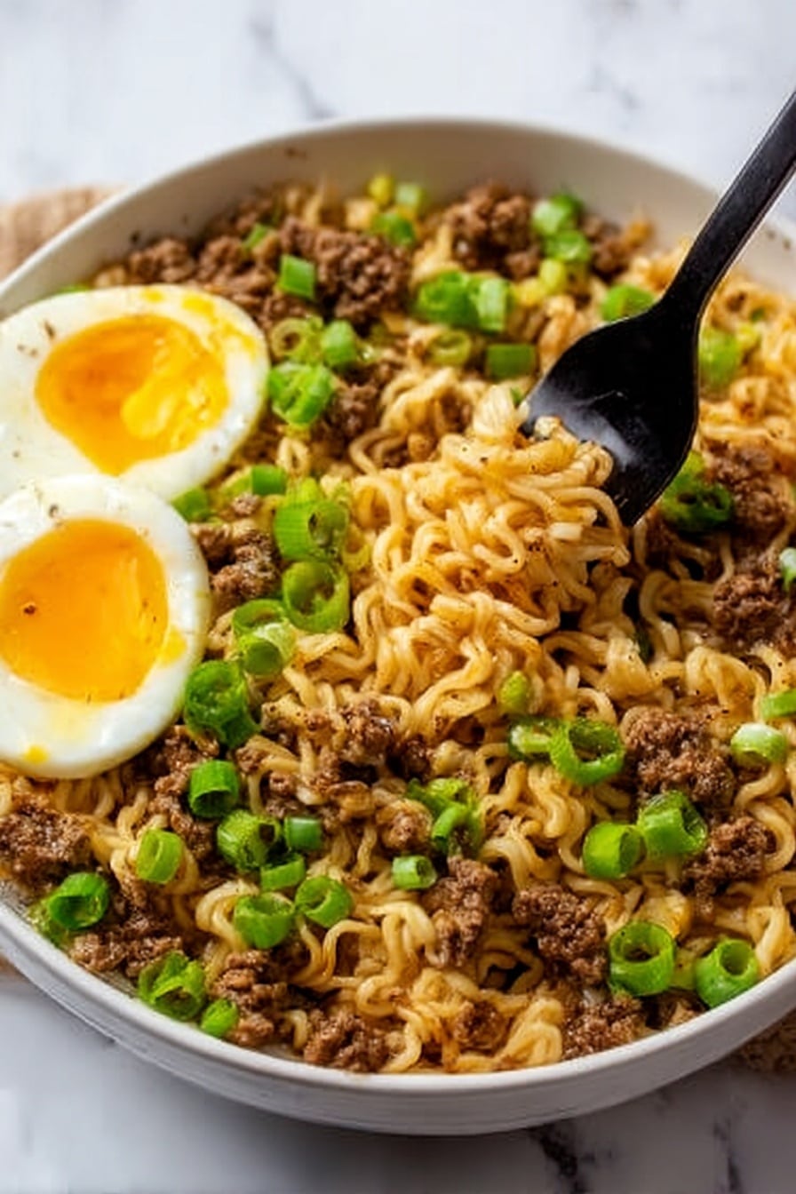 The image shows a white bowl filled with cooked ramen noodles mixed with browned ground meat and small pieces of scrambled egg. Bright green chopped scallions are sprinkled generously on top, adding fresh color. On one side of the bowl, two halves of a soft-boiled egg with a slightly runny yolk rest on the noodles. A black fork is lifting a portion of the noodles from the bowl. The bowl sits on a white marbled surface. photo taken with an iphone --ar 2:3 --v 7 - Spicy Beef Ramen, spicy ramen recipe, beef ramen bowl, easy beef ramen, flavorful ramen dish