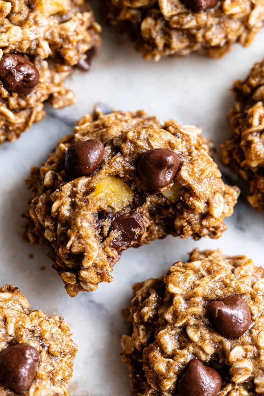 The image shows several oatmeal cookies with chocolate chips on a white marbled surface. Each cookie has a rough, chunky texture with visible oats and dark brown chocolate chips scattered throughout. One cookie is broken in half, revealing its soft, slightly moist inside with melted chocolate pieces. The cookies are light brown with some golden yellow from small bits of banana or similar fruit inside. The focus is close up, highlighting the crumbly and dense nature of the cookies. Photo taken with an iphone --ar 2:3 --v 7 - Healthy Banana Oatmeal Cookies, healthy banana cookies, easy healthy snack recipes, guilt-free dessert ideas, quick healthy cookies
