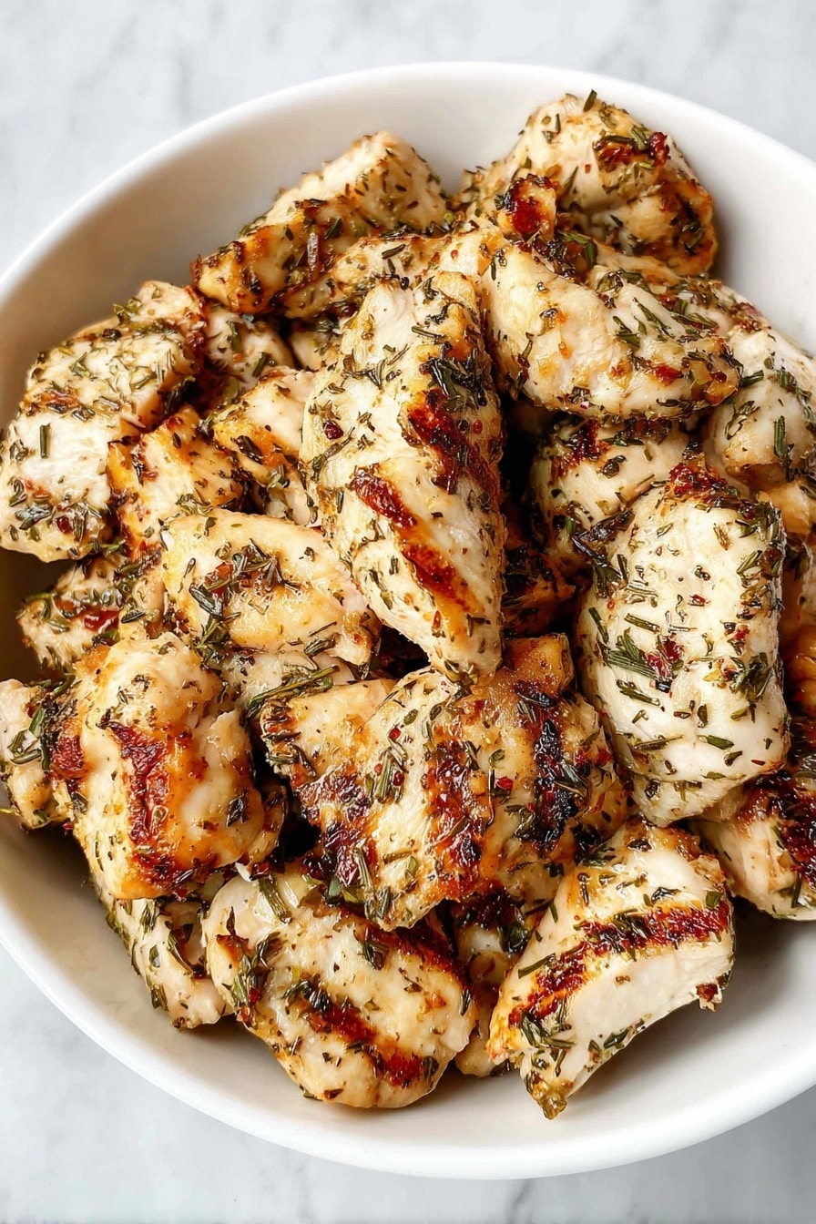 A bowl filled with cooked pieces of chicken that are seasoned with herbs and spices, each piece showing a mix of golden brown grill marks and white cooked meat with specks of green and red herbs all over. The chicken pieces are roughly shaped and layered casually in the bowl. The bowl is white and placed on a surface with a white marbled texture. photo taken with an iphone --ar 2:3 --v 7 - Copycat Chick-fil-A Grilled Chicken Nuggets, homemade grilled chicken nuggets, Chick-fil-A-inspired grilled chicken, easy grilled chicken recipes, healthy chicken nuggets