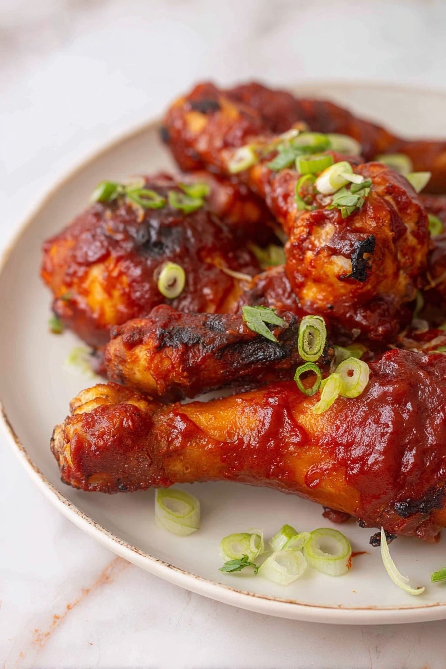 The image shows a white plate filled with five pieces of chicken drumsticks covered in thick, shiny red barbecue sauce. The chicken skin has a slightly crispy texture with some charred spots, indicating it is cooked well. On top of the chicken, there are small slices of fresh green onions scattered evenly, adding a fresh green color contrast. The plate rests on a white marbled surface that gives a clean and bright look. The photo is taken close up focusing on the front drumstick with a soft background blur photo taken with an iphone --ar 2:3 --v 7 - Baked BBQ Chicken Drumsticks, BBQ Chicken Drumsticks, Homemade BBQ Chicken, Juicy Baked Chicken Drumsticks, Easy Chicken Dinner