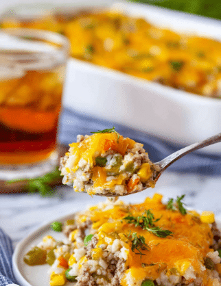 A close-up photo shows a spoon holding a scoop of a mixed dish with visible layers including yellow melted cheese, small pieces of light brown ground meat, white rice, green peas, and corn kernels, all mixed together in a creamy texture. On a white plate below the spoon, there is a serving of this dish with orange melted cheese on top and scattered green pieces, likely herbs or vegetables. In the background, there is a white baking dish filled with more of the dish topped with golden melted cheese, placed on a white marbled surface. A glass of iced tea and some green garnish are also visible behind the plate. photo taken with an iphone --ar 2:3 --v 7 - Cheesy Ground Beef and Rice Casserole, ground beef casserole recipes, easy cheesy beef rice dish, hearty comfort food dinner, quick weeknight casserole