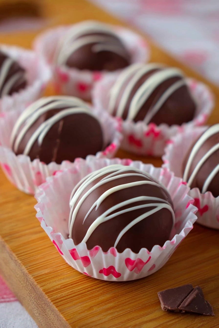 This image shows a wooden board with smooth round dark chocolate truffles placed in white paper cups with pink and red hearts design, each truffle drizzled with thin white chocolate lines on top in irregular patterns. The truffles have a shiny dark brown surface and sit in a single layer in their cups. The soft pink color of the paper cups contrasts gently with the rich chocolate, making each piece look delicate and inviting. Small pieces of chocolate can be seen around the cups. The photo taken with an iphone --ar 2:3 --v 7 - Strawberry White Chocolate Truffles, strawberry white chocolate truffles, fruit-infused chocolate truffles, easy gourmet truffles, homemade strawberry chocolates
