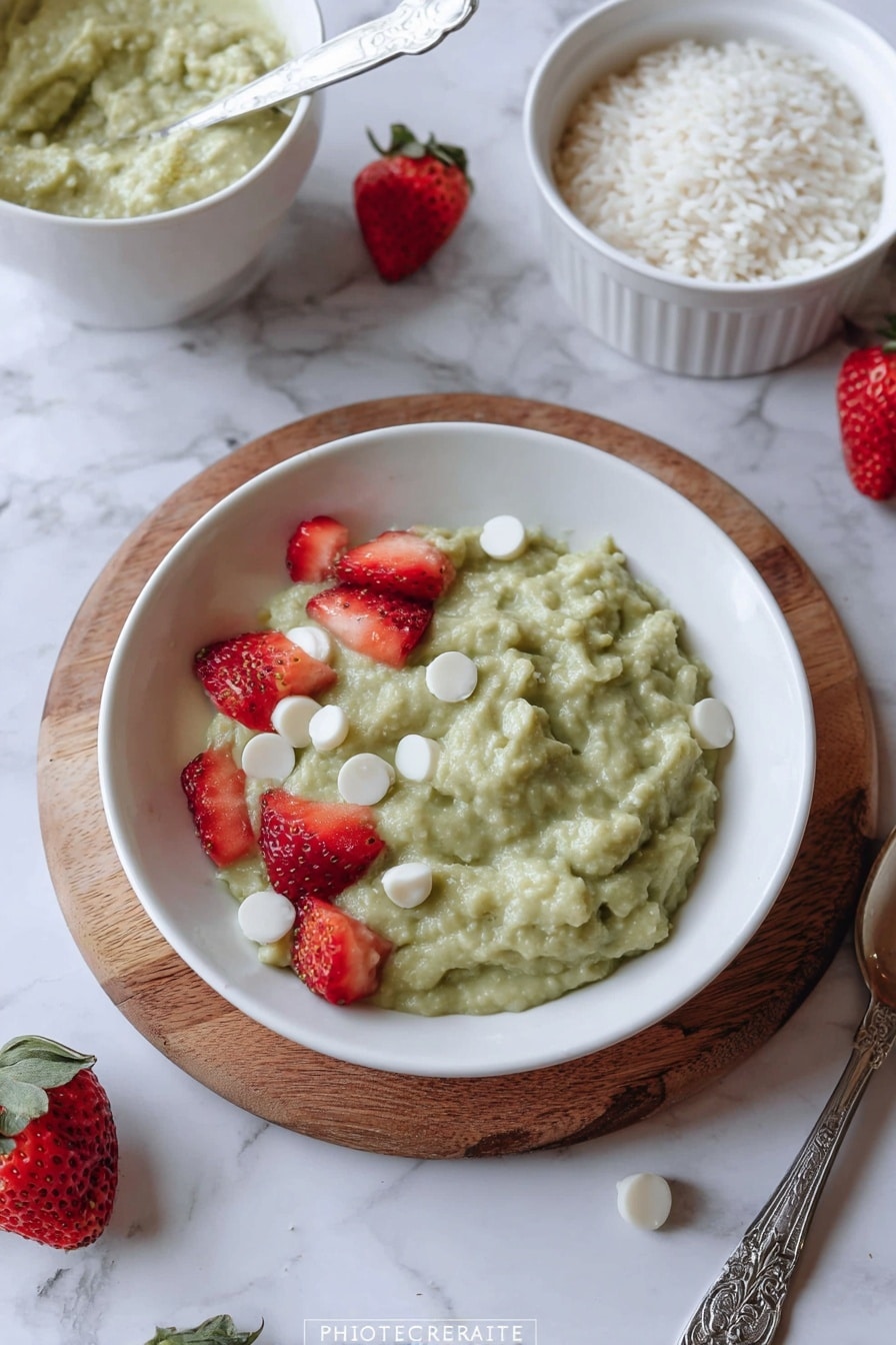 A white plate sits on a wooden board with a green creamy mixture that has a chunky texture, topped with small white discs and pieces of red strawberries scattered on top. Around the plate on a white marbled surface are a few whole strawberries, a white bowl filled with the same green creamy mixture with a spoon inside, and another white bowl with white rice grains. A silver spoon handle with a decorative pattern lies next to the plate. photo taken with an iphone --ar 2:3 --v 7 - Matcha Rice Pudding, matcha dessert recipes, green tea pudding, easy rice pudding, comforting matcha treat