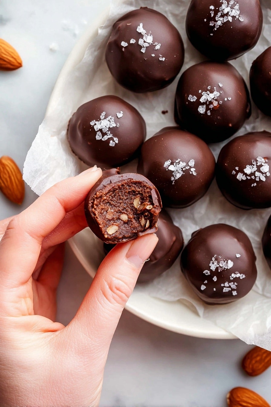 The image shows a woman's hand holding a round chocolate truffle with a bite taken out, revealing a dense, dark brown filling with small bits inside. Around the hand, there are multiple whole chocolate truffles, smooth and shiny with a dark brown color, each topped with a few flakes of sea salt. Whole almonds in rich brown tones are scattered among the truffles. The truffles are arranged on white parchment paper inside a white plate, set on a white marbled surface. photo taken with an iphone --ar 2:3 --v 7 - Healthy Chocolate Date Almond Truffles, healthy chocolate truffles, guilt-free dessert recipes, natural ingredient truffles, homemade healthy treats