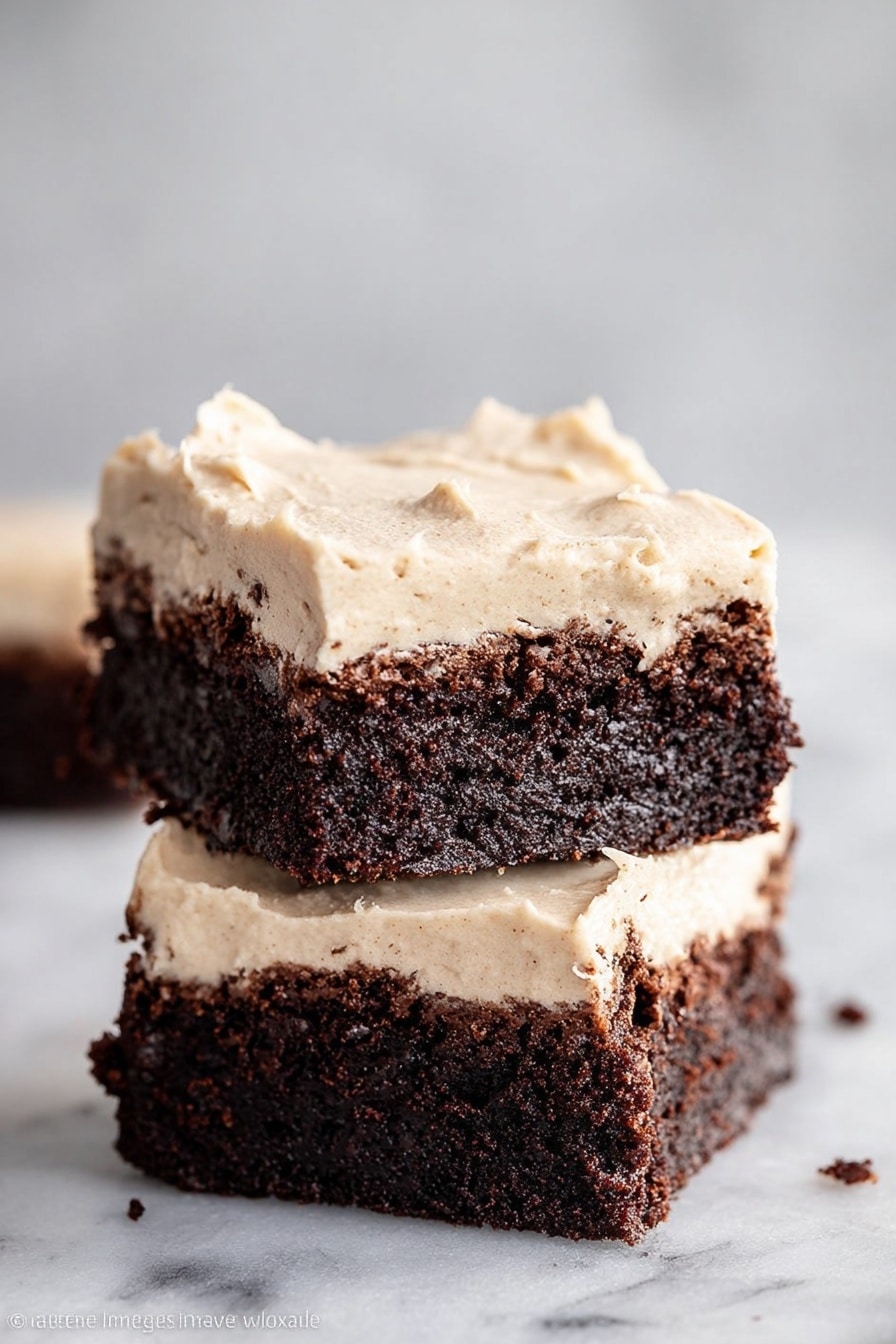 Two square pieces of dark brown chocolate brownie are stacked on top of each other against a soft, light gray background. Each brownie has two layers: the bottom layer is thick and fudgy with a rough, crumbly texture, while the top layer is a smooth, creamy, light beige frosting spread unevenly with some peaks and swirls. Small crumbs are visible on the surface, adding a natural look. The brownies sit on a white marbled surface. photo taken with an iphone --ar 2:3 --v 7 - Guinness Chocolate Brownies with Baileys Frosting, Guinness Brownies recipe, Baileys frosting, fudgy Guinness brownies, chocolate dessert with Baileys