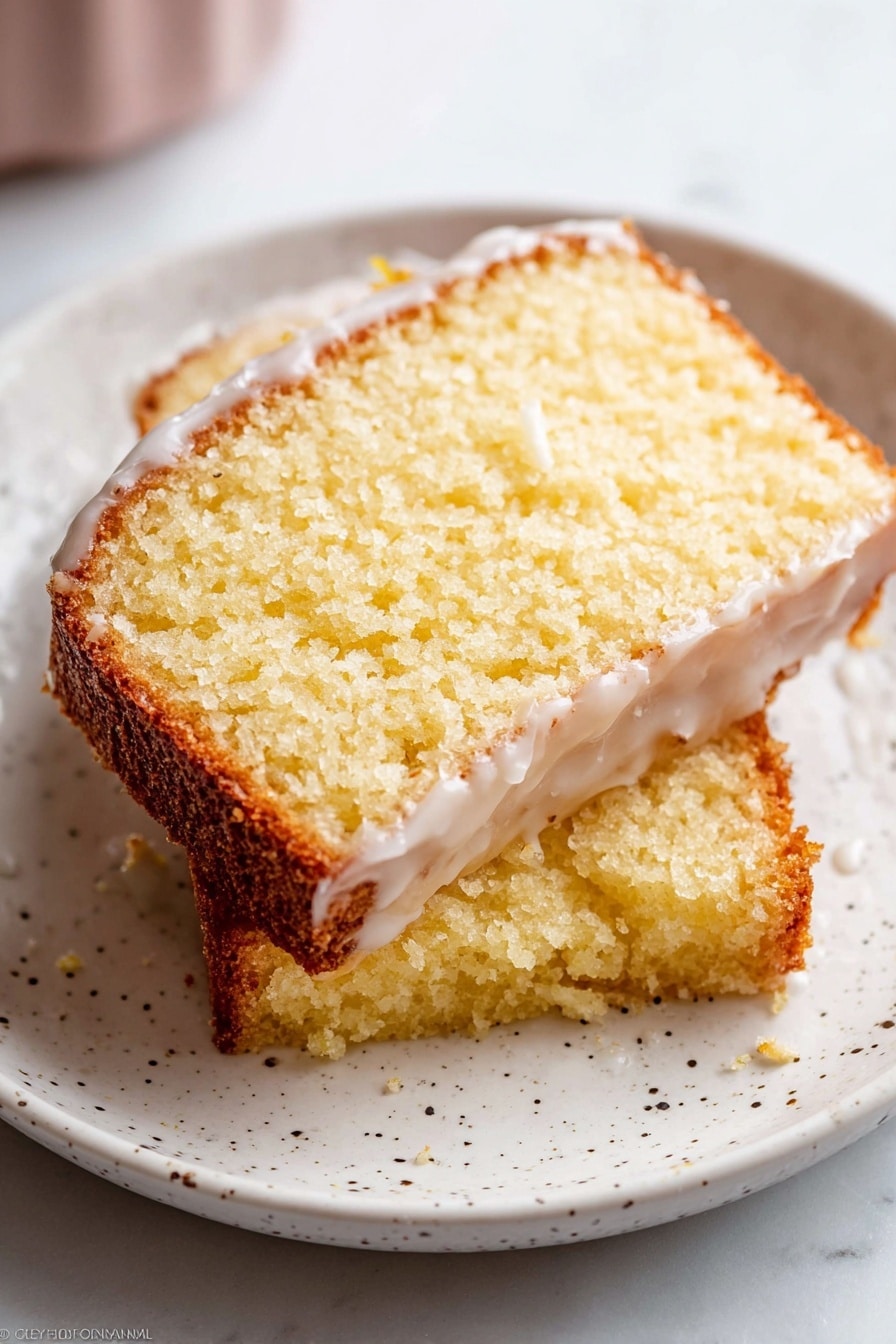 Two slices of yellow cake with a light texture and crumbly edges are stacked on a white plate with small black specks. The top slice has a thin layer of white glaze that partly covers the surface, showing a moist interior. The cake edges are golden brown, with a soft, spongy middle layer that is pale yellow. The plate sits on a white marbled surface, and the image has soft natural light highlighting the texture of the cake. photo taken with an iphone --ar 2:3 --v 7 - Iced Lemon Pound Cake, Lemon Pound Cake, Lemon Cake Recipe, Moist Lemon Pound Cake, Zesty Lemon Dessert