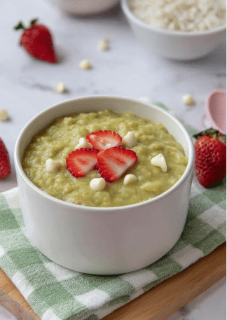 A white bowl is filled with greenish thick porridge that looks creamy and soft, topped with a few white chocolate chips and three sliced red strawberries arranged in the center. The bowl sits on a green and white checkered cloth, which is placed on a wooden board. Around the bowl, there are scattered strawberries and white chocolate chips on a white marbled surface, adding color contrast. The background shows a larger white bowl with white rice and a spoon, all in soft natural light. Photo taken with an iphone --ar 2:3 --v 7 - Matcha Rice Pudding, matcha dessert recipes, green tea pudding, easy rice pudding, comforting matcha treat